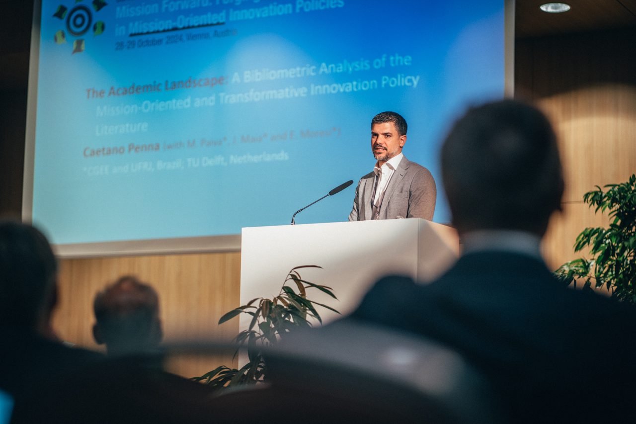 Caetano Penna speaking at a lectern in front of a projected title slide, seen past the heads of the audience